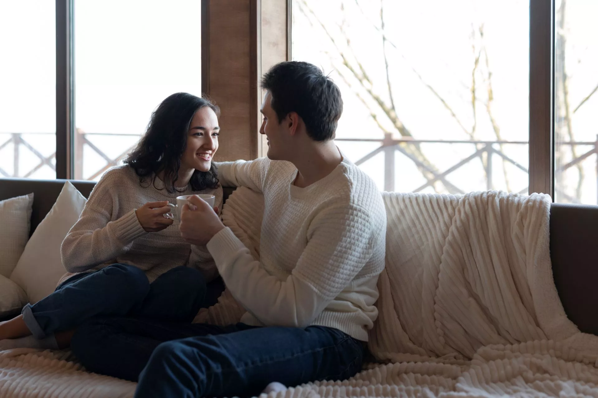 室內客廳中，一對情侶坐在沙發上對視微笑，各自手拿馬克杯聊天，窗邊自然光灑入，氛圍溫馨放鬆。