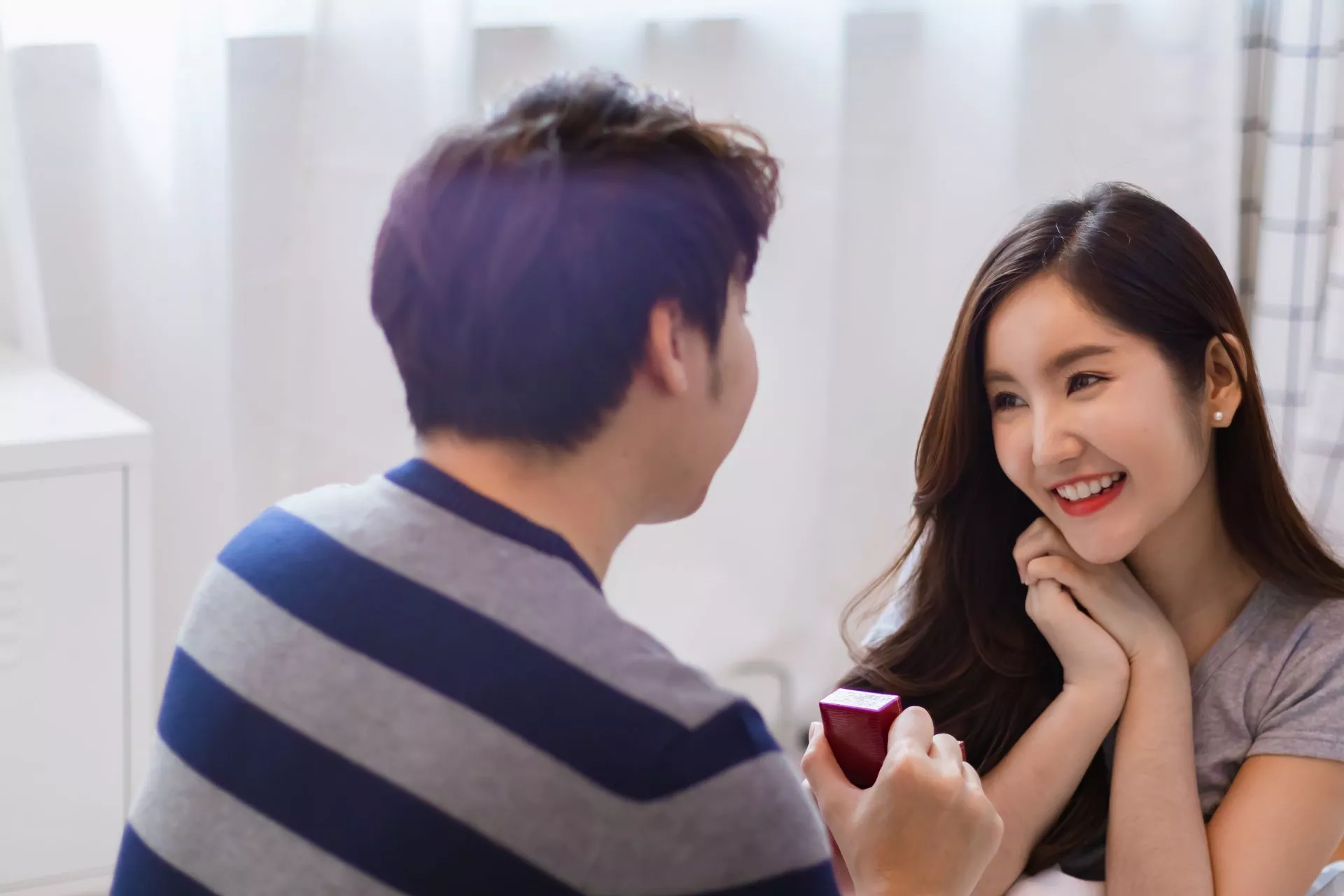 室內空間中，一名男子手持戒指盒向女子求婚，女子雙手托腮露出驚喜微笑，兩人面對面互動，氣氛溫馨浪漫。