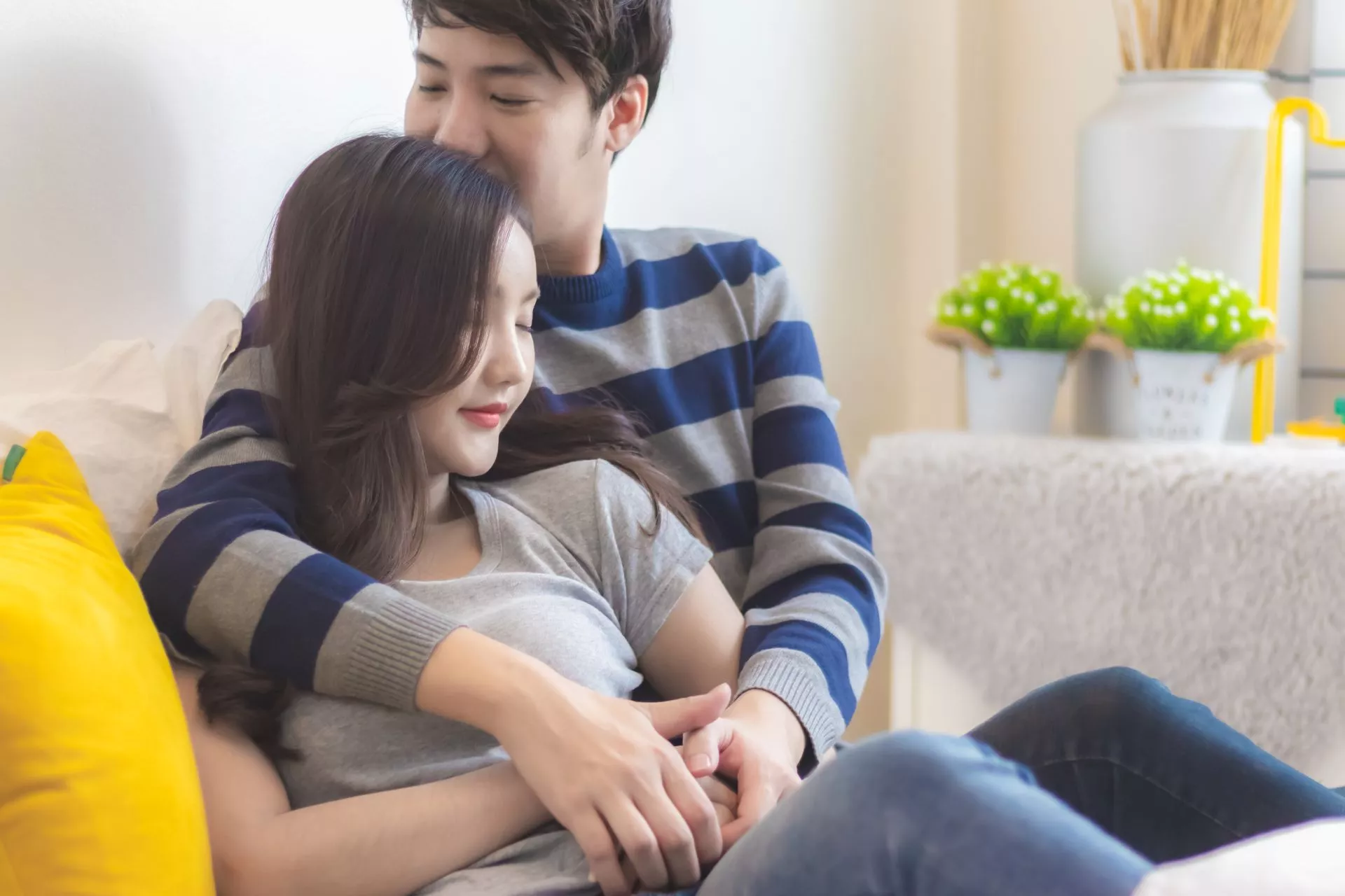 一對男女在室內沙發上相互依偎，男子從後方環抱女子，女子閉眼微笑放鬆坐在懷中，背景可見抱枕與盆栽，呈現溫馨居家相處情境。